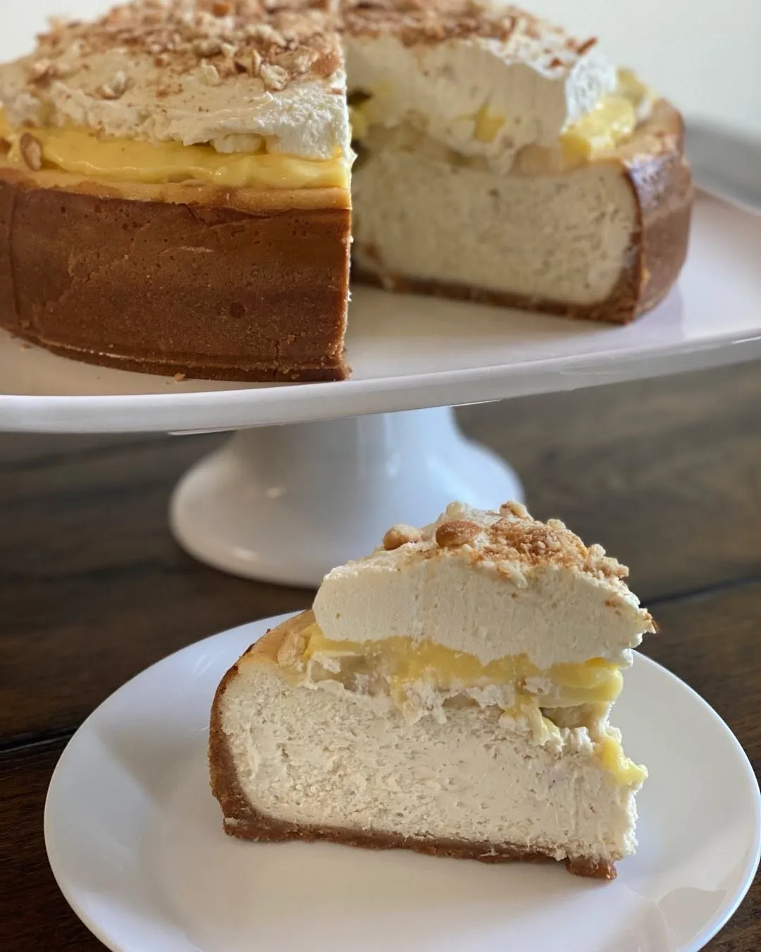 A banana pudding cheesecake with a banana-custard layer, whipped cream and crushed vanilla wafers, with a slice on a plate.