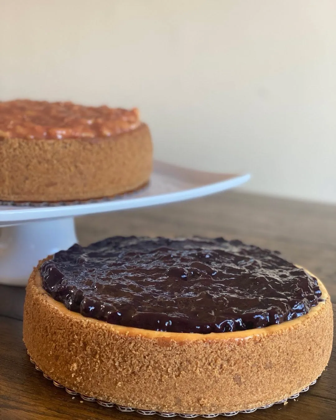 A blueberry cheesecake topped with blueberry compote, with an apple cheesecake on a stand behind.