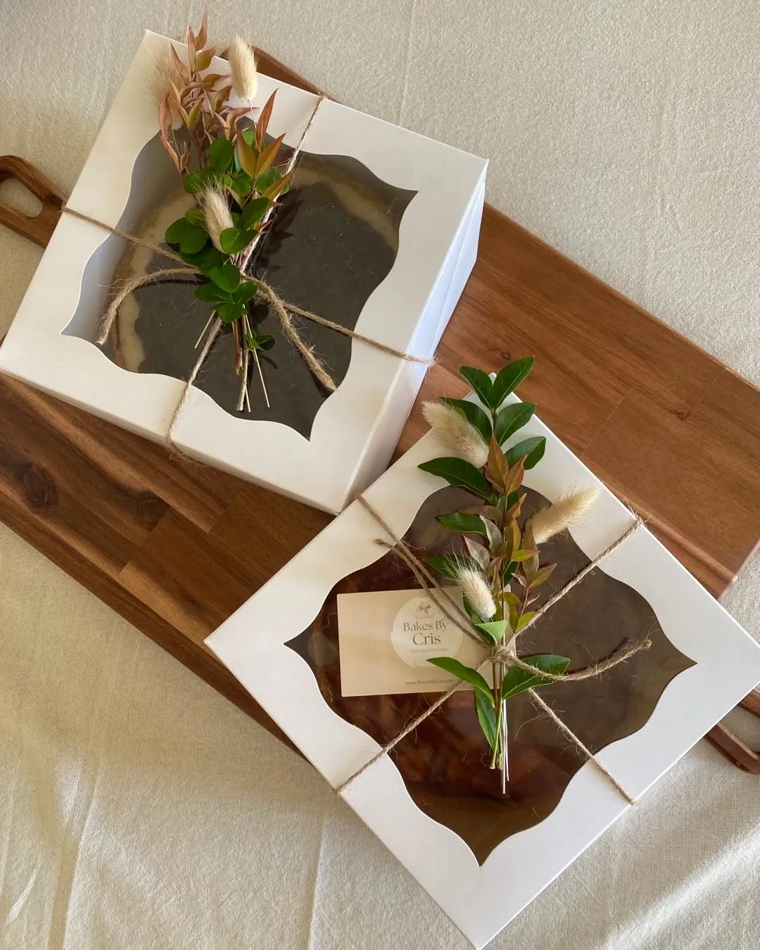 Two boxed cheesecakes with a Bakes by Cris label, decorated with greenery.
