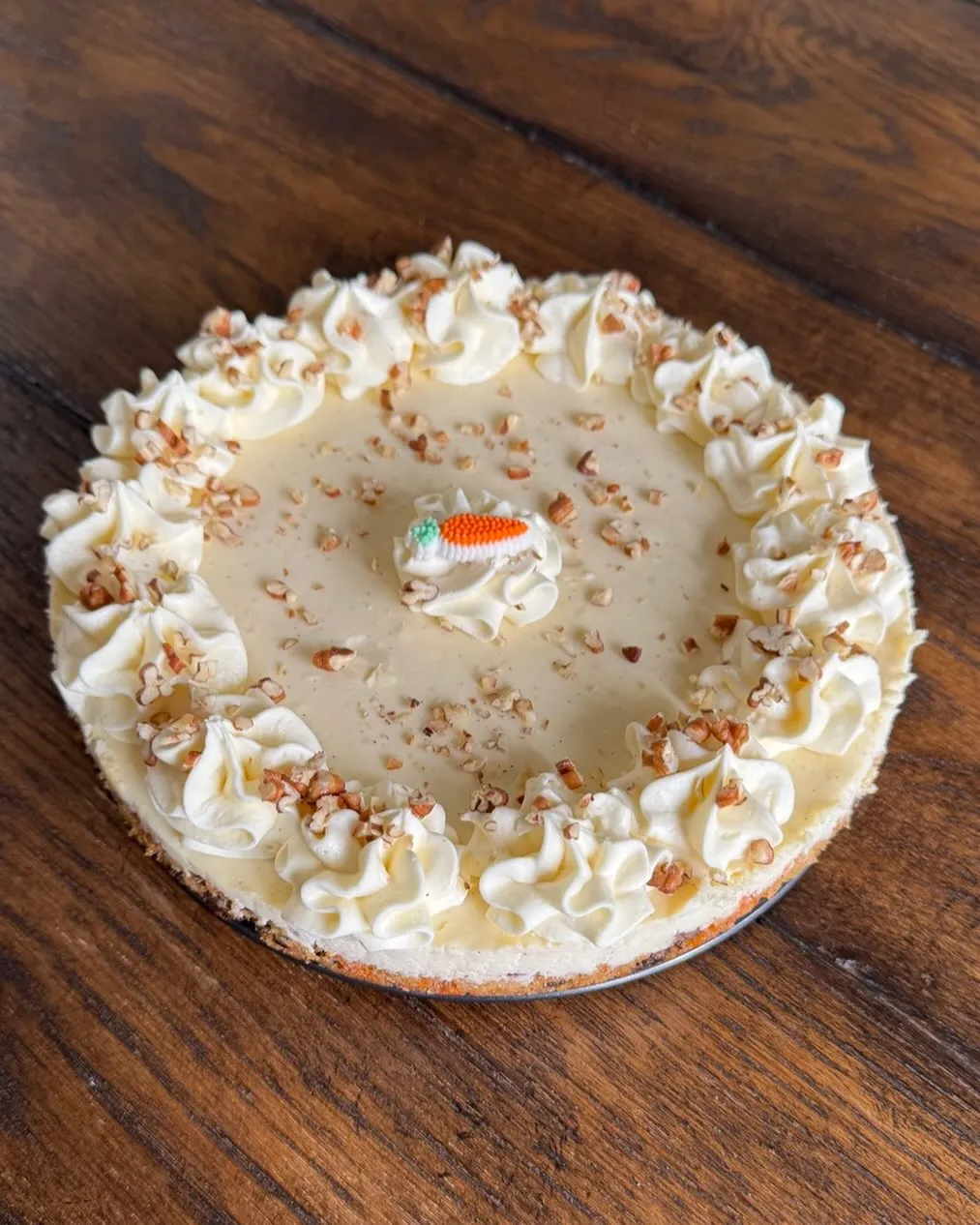A carrot cake cheesecake ringed with cream-cheese rosettes and chopped pecans, with a small marzipan carrot on top.