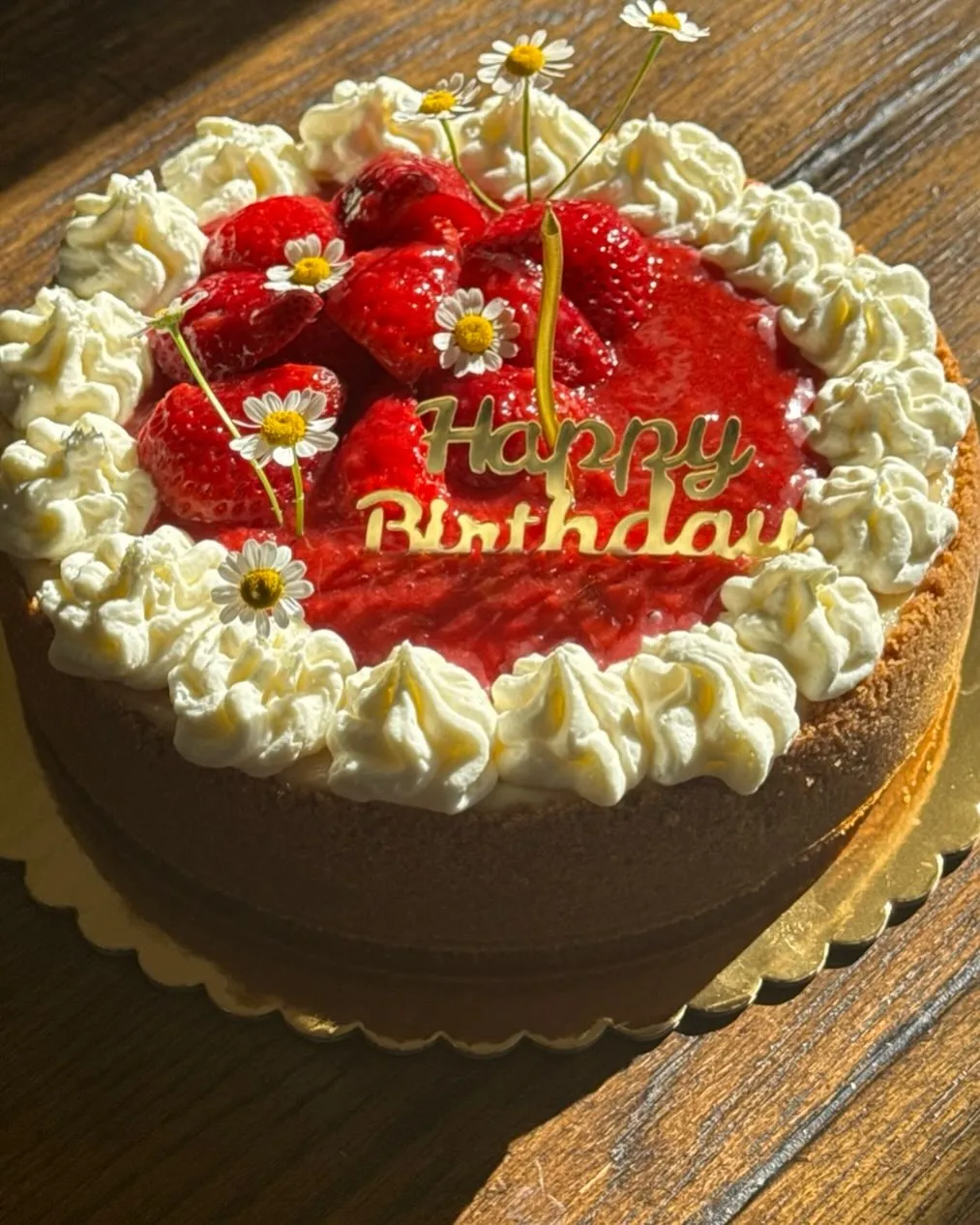 A strawberry cheesecake decorated with a gold "Happy Birthday" topper, fresh strawberries, glaze and daisies.