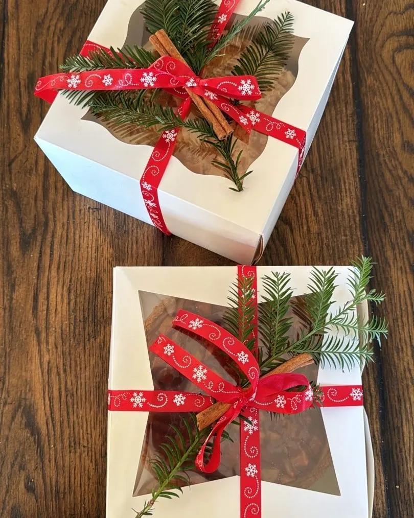 Two cheesecakes in white gift boxes tied with festive red ribbon, cinnamon sticks and pine sprigs.