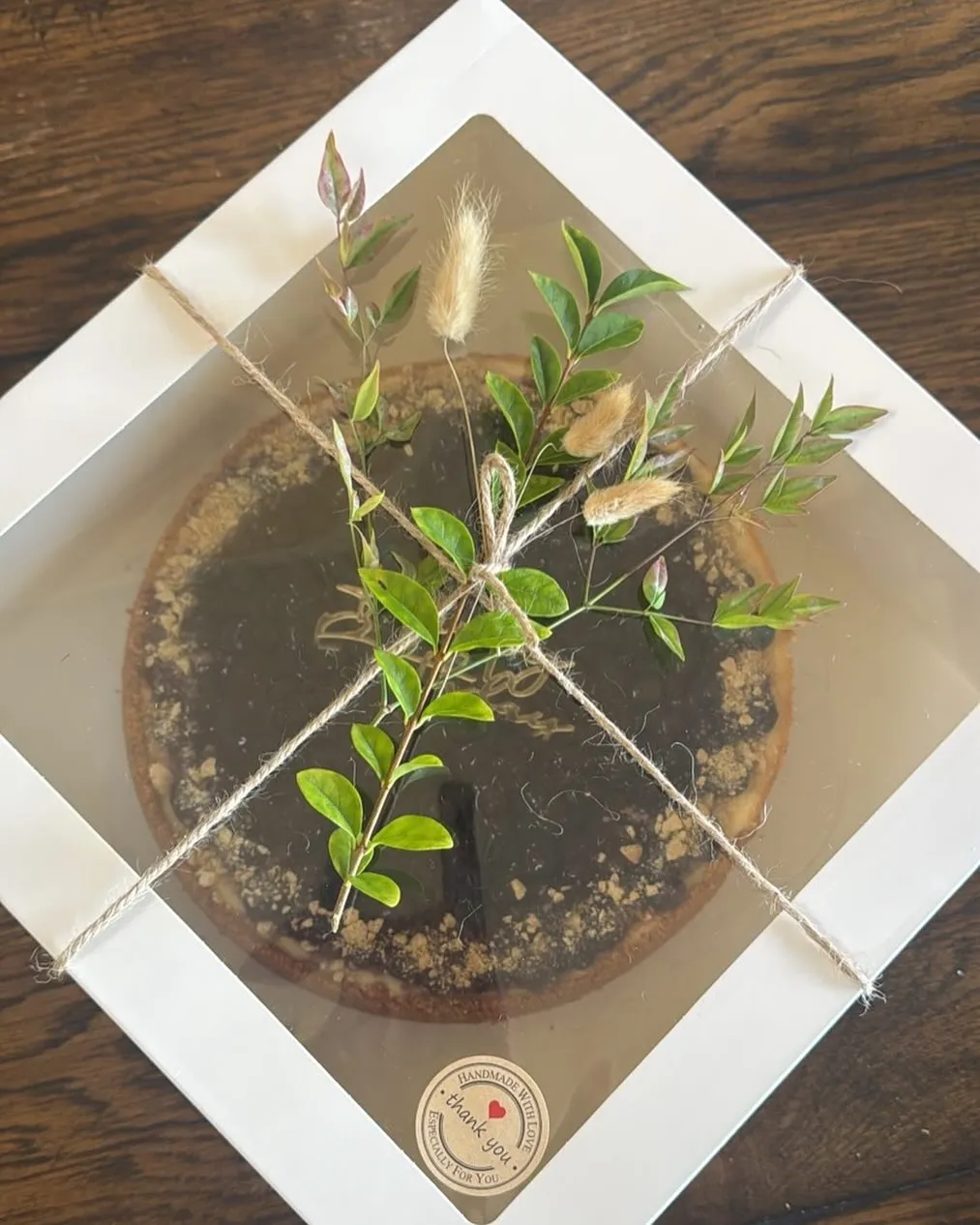 A boxed cheesecake topped with dark berry compote, decorated with greenery and a thank-you sticker.