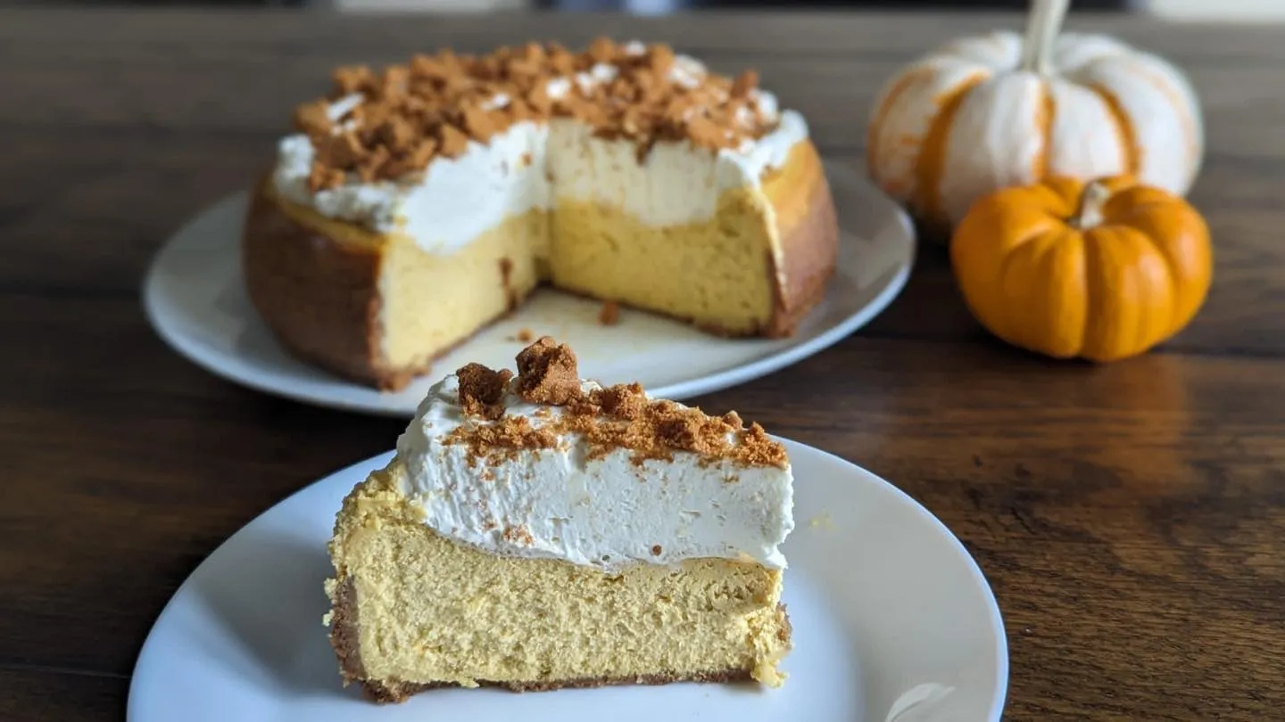 A pumpkin spice cheesecake with a Biscoff crust, topped with whipped cream and Biscoff crumble, with a slice on a plate.